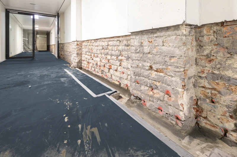 Der Putz einer Ziegelwand ist zu einem Drittel vom Fußboden her abgetragen, damit Abdichtungsmaßnahmen am denkmalgeschützten Gebäude vorgenommen werden können.